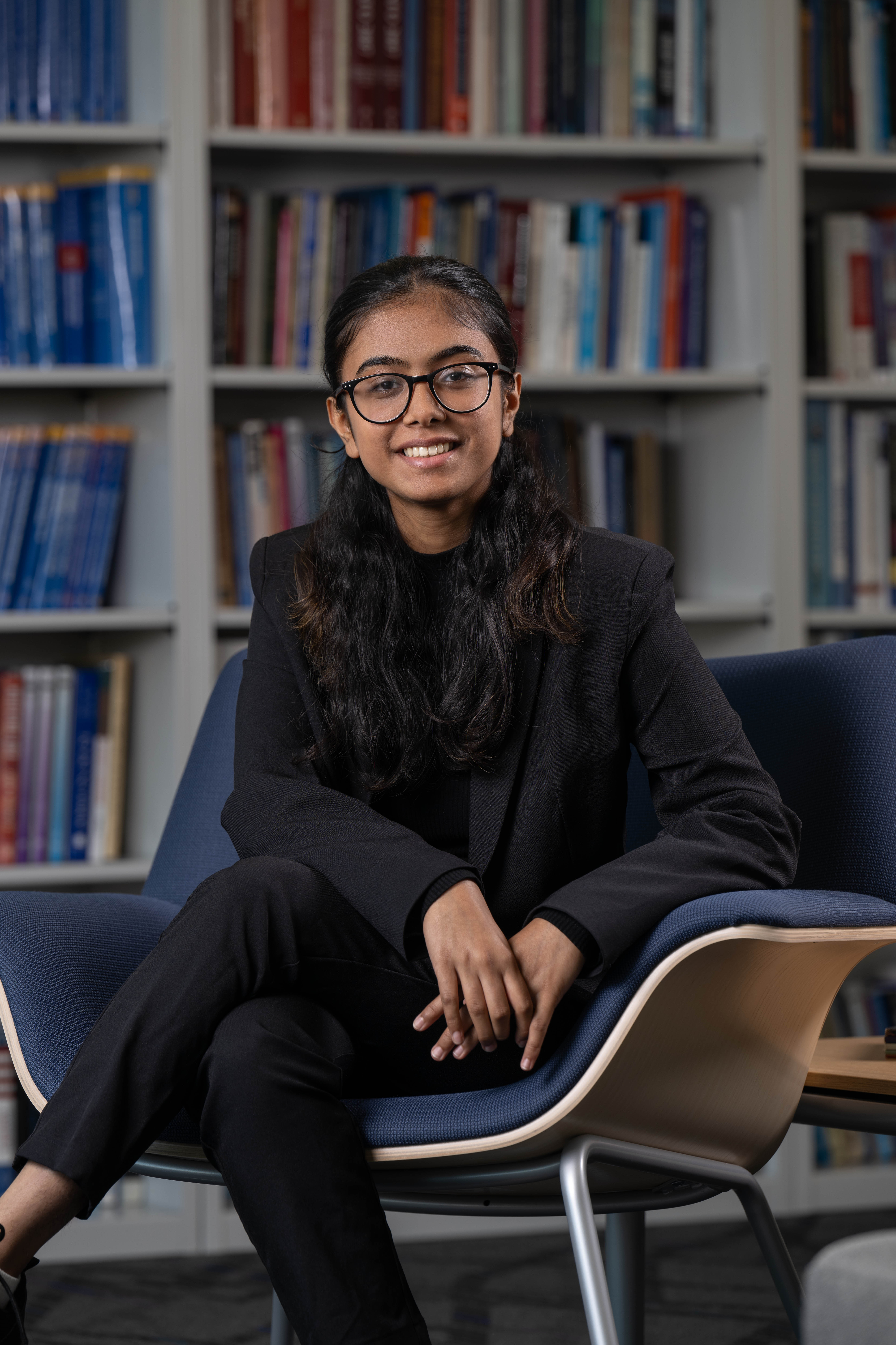 Professional portrait of a smiling young lady who appears to be South Asian with long wavy black hair and round glasses with a black frame sitting cross-legged on a blue chair. She's wearing a black blazer and pants with a blurred bookshelf in the background.