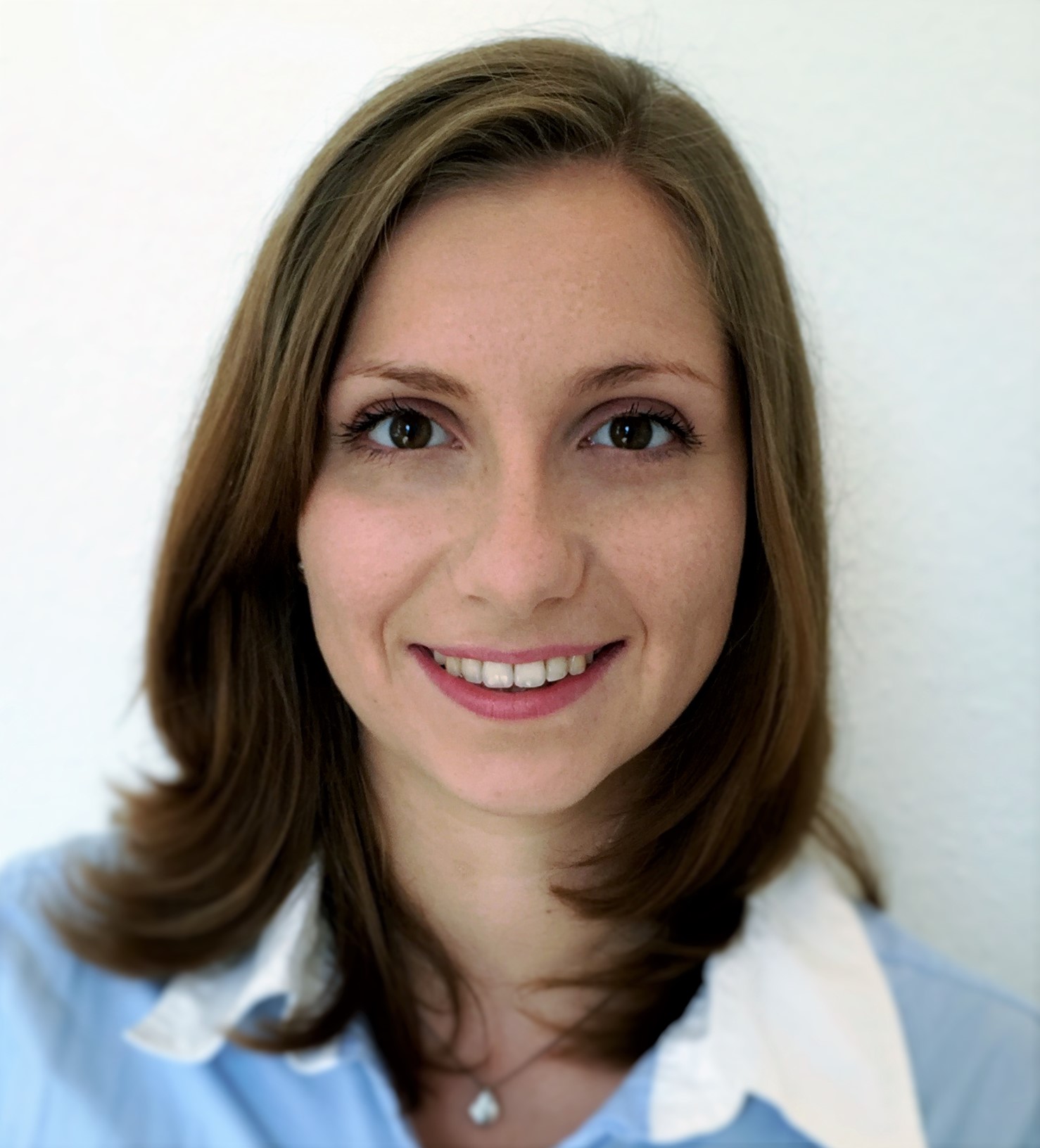 Headshot of a woman who appears to be white with long brown hair and an open bright smile, wearing a blue dress shirt with a white collar and a pearl necklace, in front of a white background.