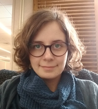Headshot of a smiling young woman who appears to be white in front of a wooden panel. She has shoulder length brown, slightly wavy hair and is wearing large round brown glasses and small circular earrings. A large blue knitted scarf is around her neck and she’s indoors wearing a sweater in a slightly different shade of blue/green.