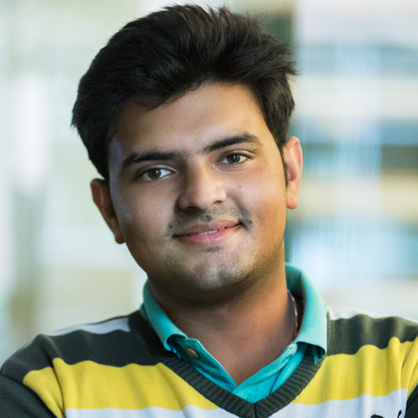 Headshot of a smiling young man with dark brown hair who appears to be Asian. He is wearing a turquoise shirt under a yellow striped v-neck sweater. The background is bright and blurred in a bokeh effect.