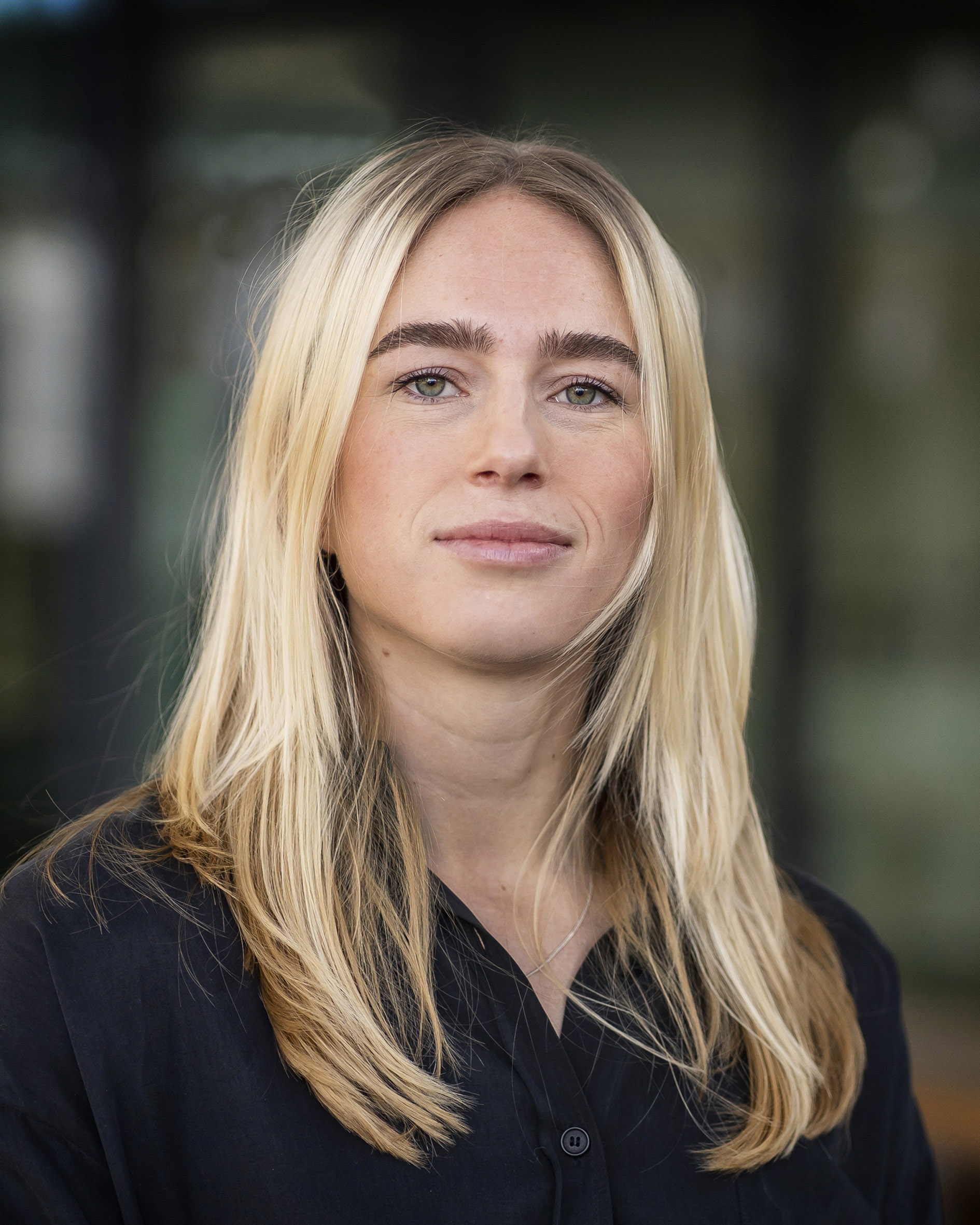 Professional headshot of a young white woman with middle long blonde hair, wearing a dark gray blouse. She’s standing outside in front of a building, the background is blurred.