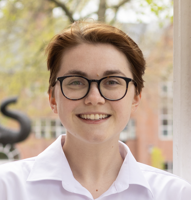 Headshot of a smiling young white person with short red hair and glasses. They are wearing a white collared shirt and standing outside of an academic building.