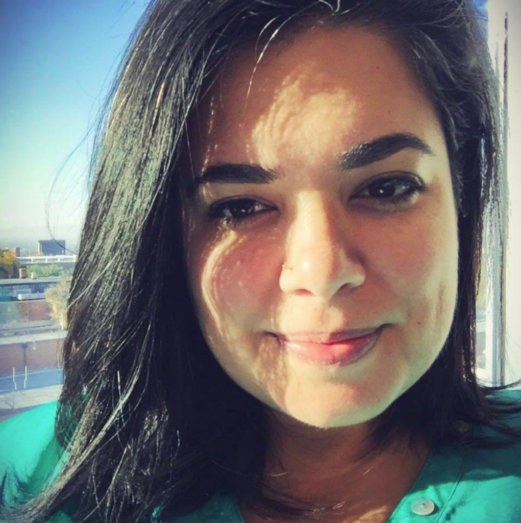 Selfie of a young woman who appears to be white in front of a window. She has long, straight, black hair, dark eyes, and is slightly smiling. She has a small golden nose piercing and is wearing a teal shirt.