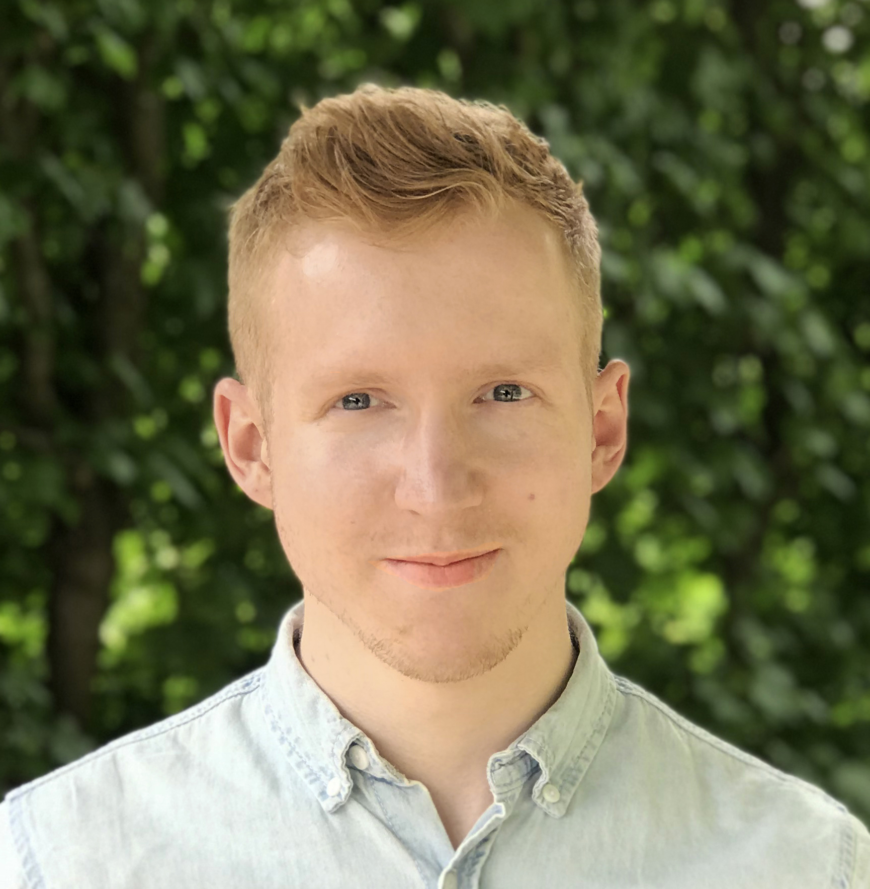 Photo of a white man with short light hair and a hint of a smile, wearing a light blue shirt. He’s standing in front of a green background, indicating he’s outside.