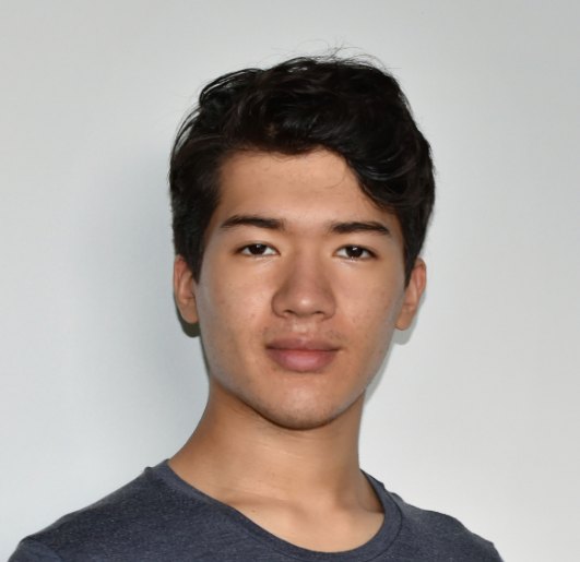 Headshot of a young man with medium tone skin, and short brown hair offering a slight smile wearing a blue tee shirt in front of a solid-colored light background.