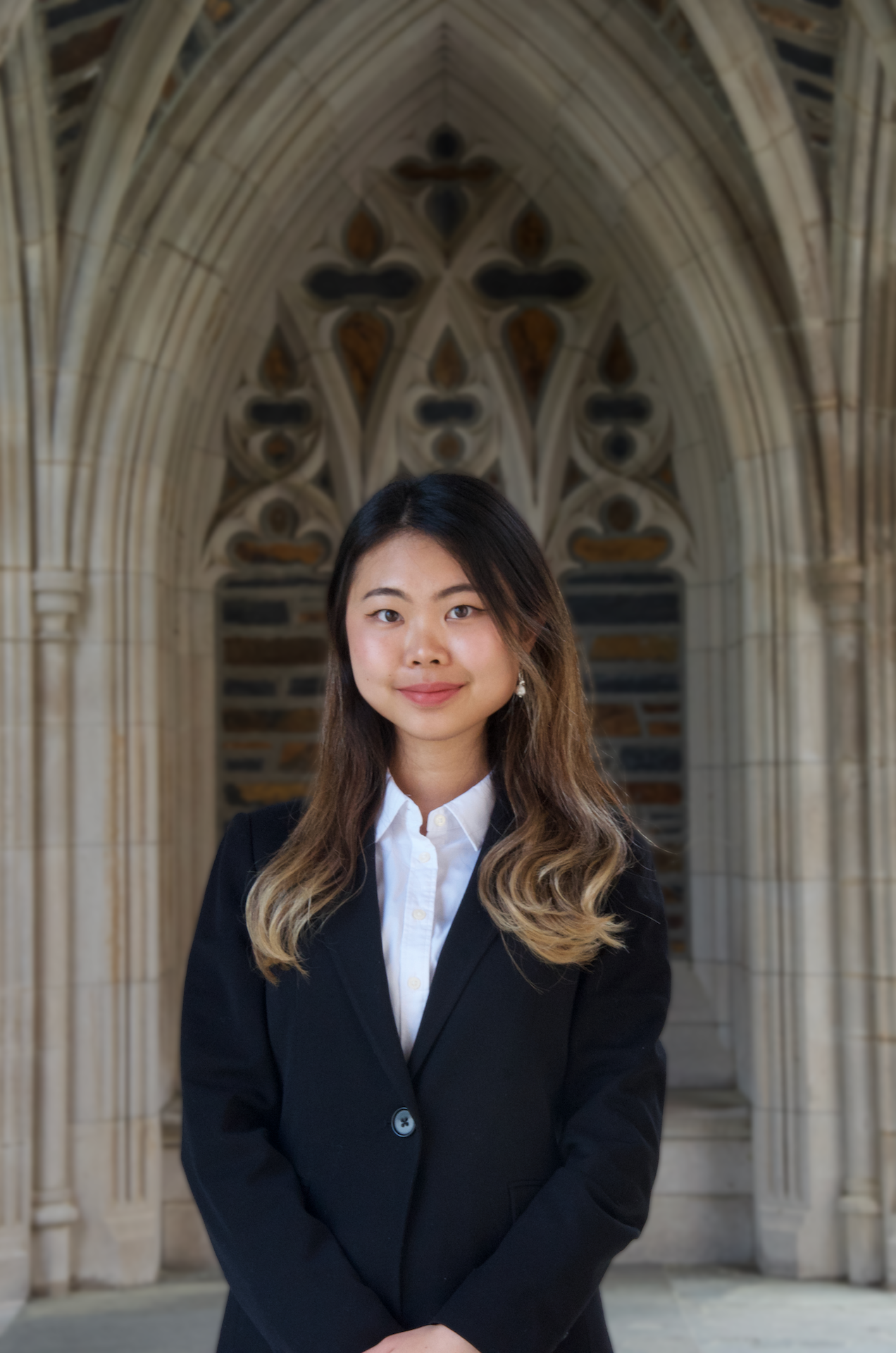 Professional headshot of a smiling young East Asian woman with long wavy hair in brown-blonde ombré hues. She is wearing a black blazer and a white collared shirt, with gothic architecture in the background.
