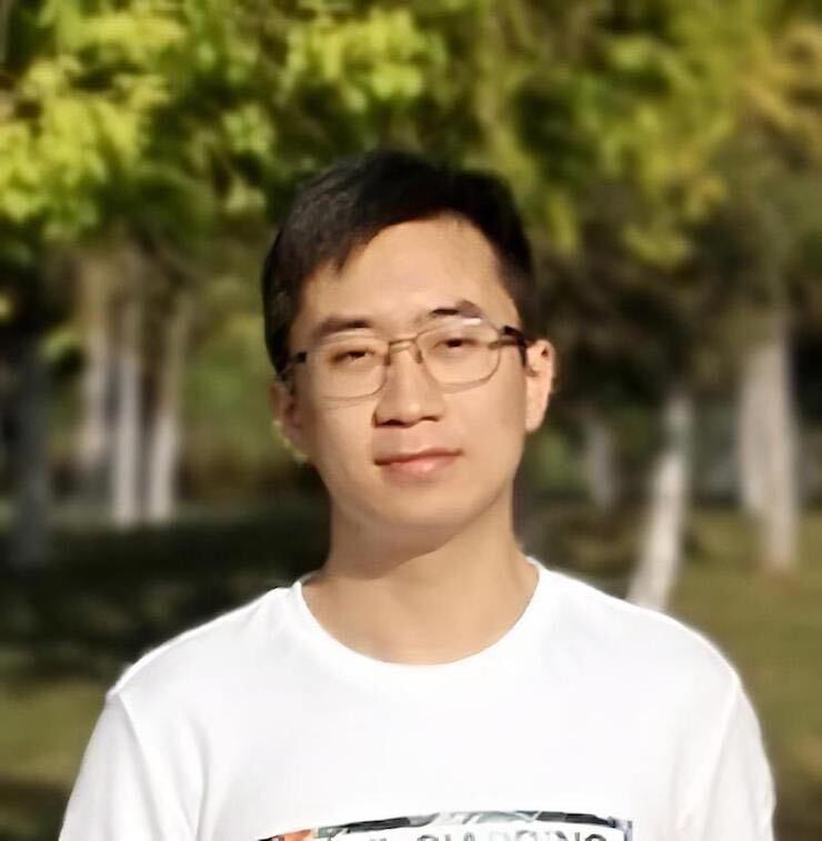 Photo of a young man with short dark hair who appears to be Asian. He’s wearing glasses with square lenses and a white graphic T-shirt and standing outside with trees seen in the background.