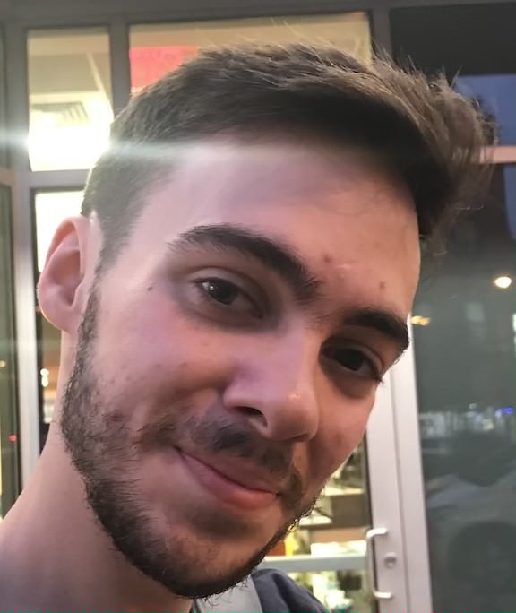 Photo of a smiling young man with dark brown hair who appears to be white. He has brown eyes and stubble, and is standing in front of a door.