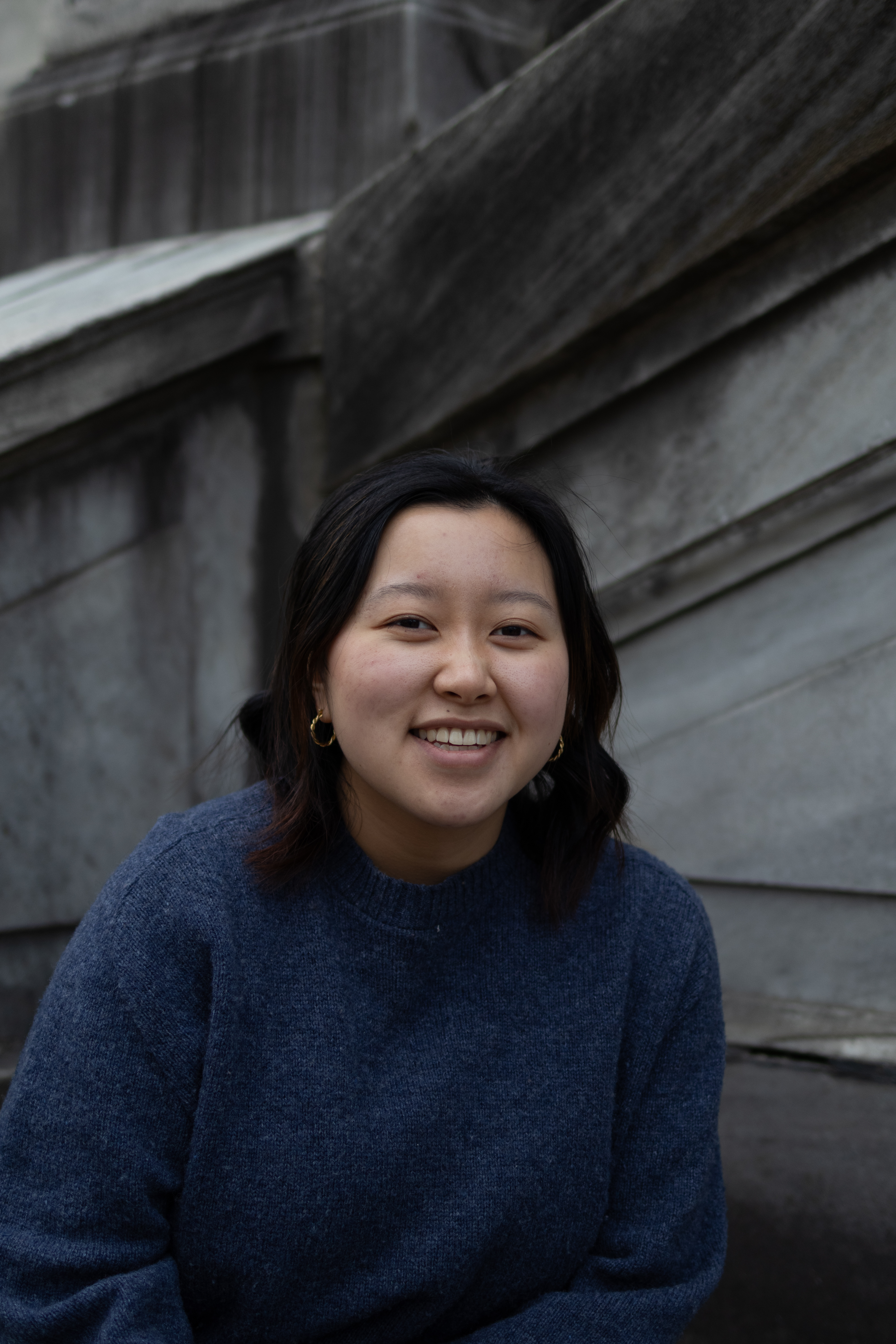 Photograph of a young Asian woman with shoulder length black hair. She smiles widely while wearing a dark blue sweater in a grey marble background.