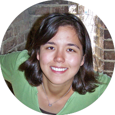 Headshot of a young, light-skinned woman taken in front of a brick wall. She has dark, medium-long hair, dark eyes and has a bright-white smile. She’s wearing a green t-shirt and has a small silver ring on a necklace around her neck.