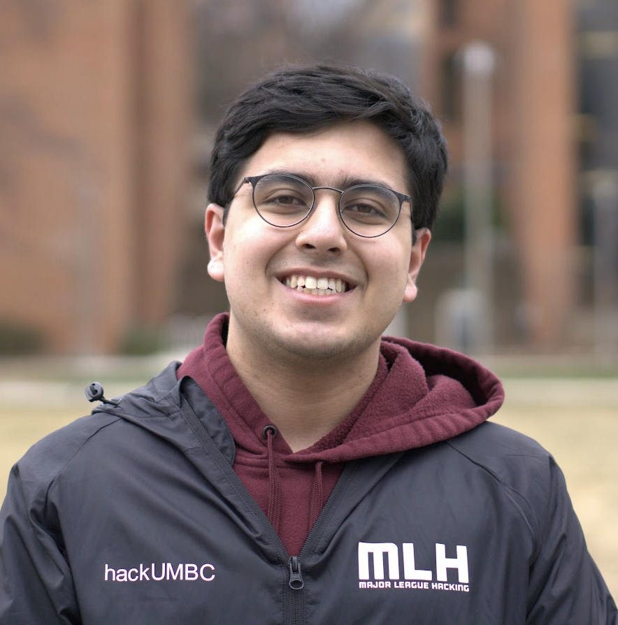 Headshot of a young man who appears to be Asian with wavy dark hair and round glasses wearing a maroon hoodie and black jacket embroidered with “hackUMBC” and “Major League Hacking”. He is standing outside in front of a brick building, and the background is blurred.