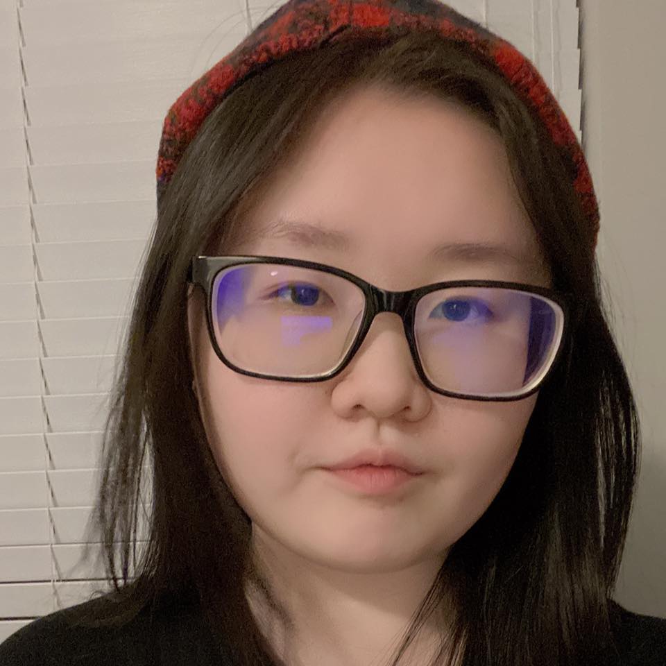 Closeup photo of a young female who appears to be Asian with dark hair wearing a plaid beanie and black glasses. The photo is taken inside, on the background there are window blinds.