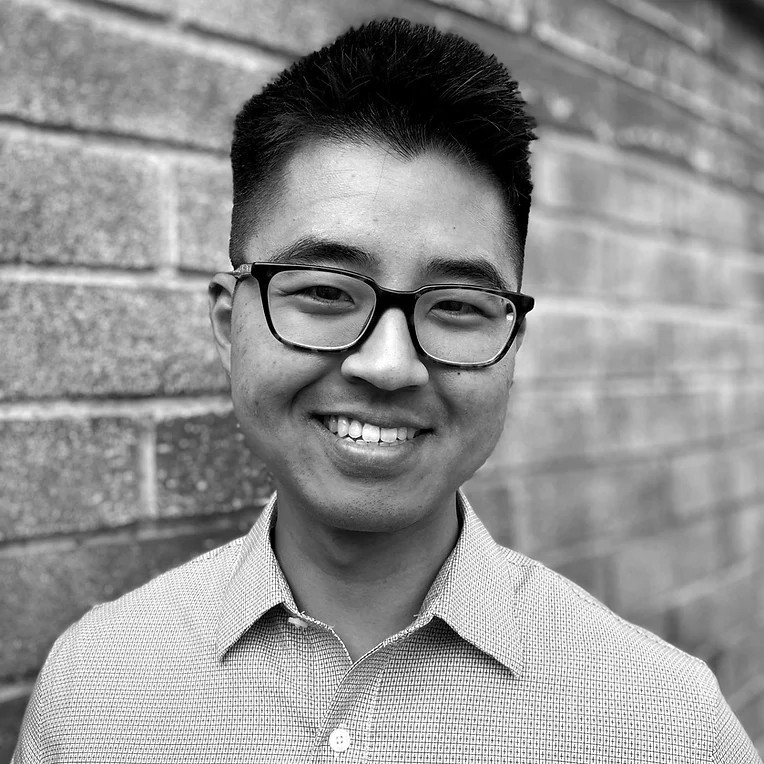 A black and white photo of a young male who appears to be Asian with a full smile. The person is wearing dark-colored frame glasses and a light-color collar button-down shirt and is next to a brick wall.