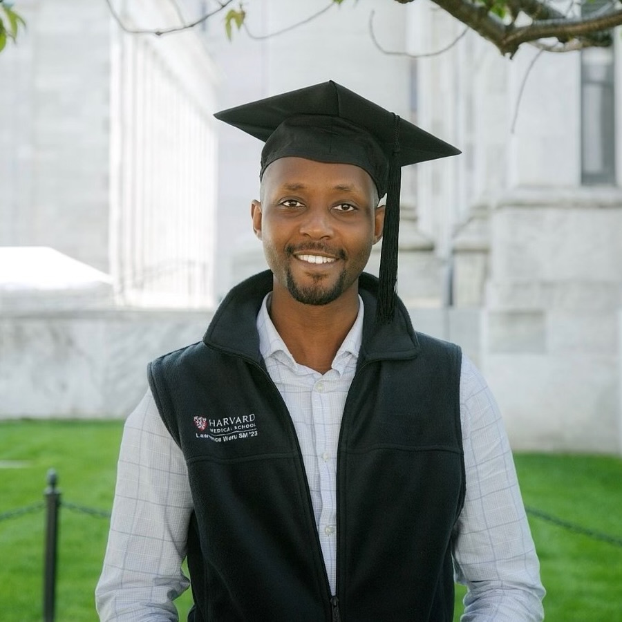 Photograph of a young black male with a full smile framed by a goatee. He wears a black fleece vest over a white checkered dress shirt and a graduation cap. He stands in front of the fenced grass of a marble building.