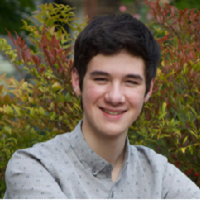 Photo of a smiling young man who appears to be Asian with wavy dark hair wearing a gray collared shirt. He is in front of green and red foliage.