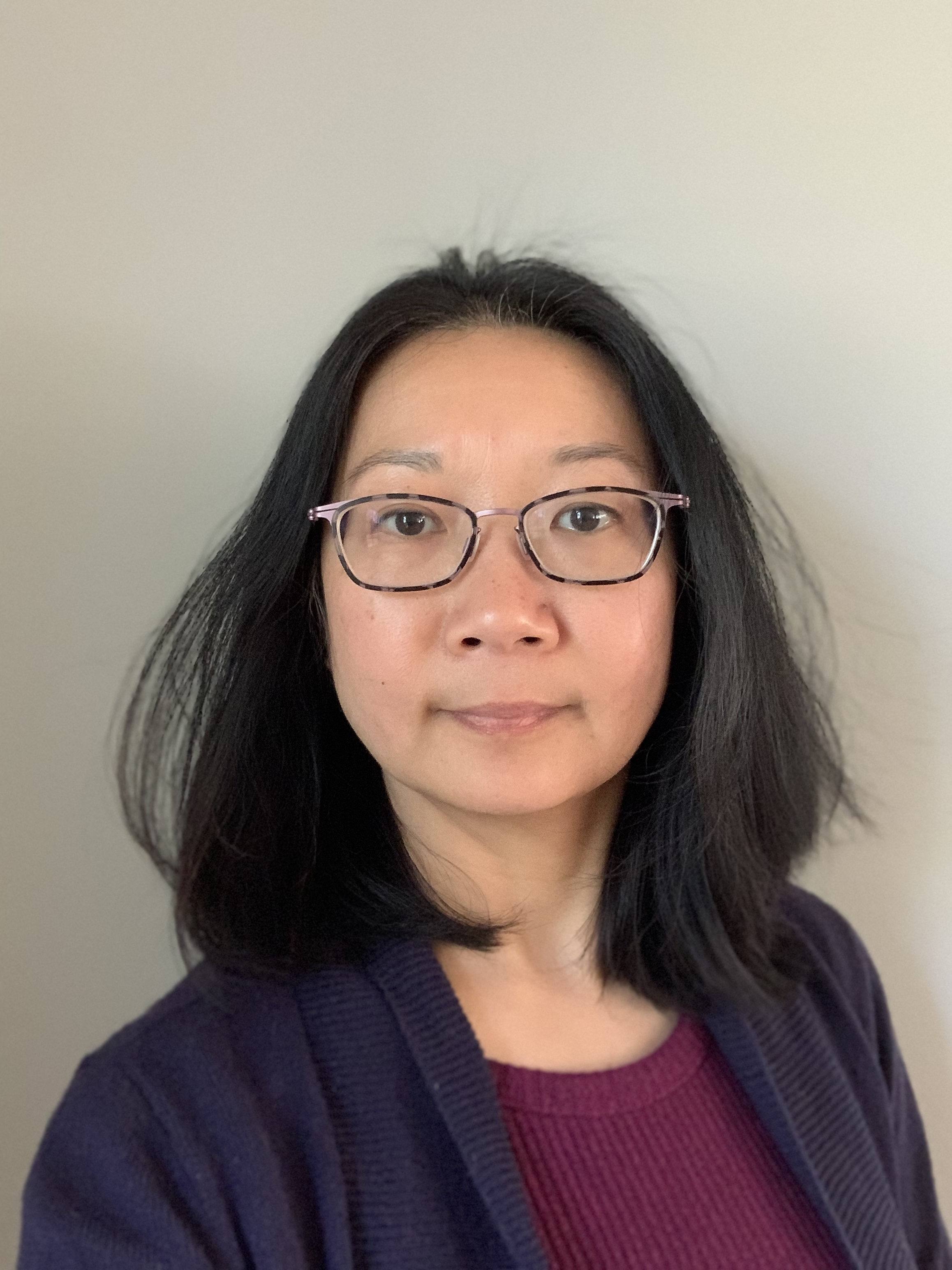 Photograph of an Asian female with shoulder length black hair wearing thin tortoiseshell glasses and a navy cardigan over a maroon sweater in front of a solid-colored light background.