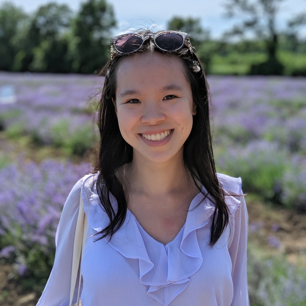 Photo of a smiling young woman who appears to be Asian with dark hair wearing a lavender shirt. There are sunglasses resting on her head and she is in front of a field of purple flowers on a sunny day.