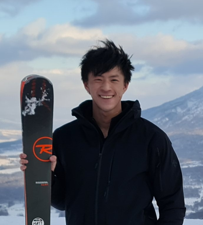 This photo shows a young man who appears to be Asian on top of a snowy mountain holding a pair of skis. He’s got medium-short dark hair and is smiling broadly. He’s wearing a black winter jacket.