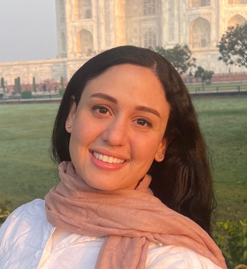 Photo of a smiling young woman with dark hair and brown eyes wearing a white shirt and a peach-colored scarf. She is standing in front of an ornate building and garden with a cloudy sky.