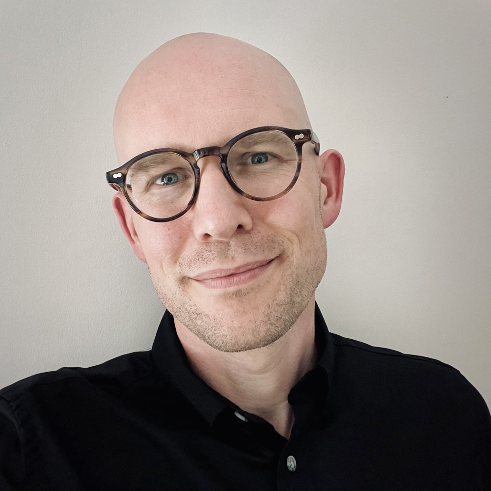 Headshot of a smiling middle-aged white man with a shaved head and round glasses with brown frames. He is wearing a black button down shirt in front of a solid light-colored background.