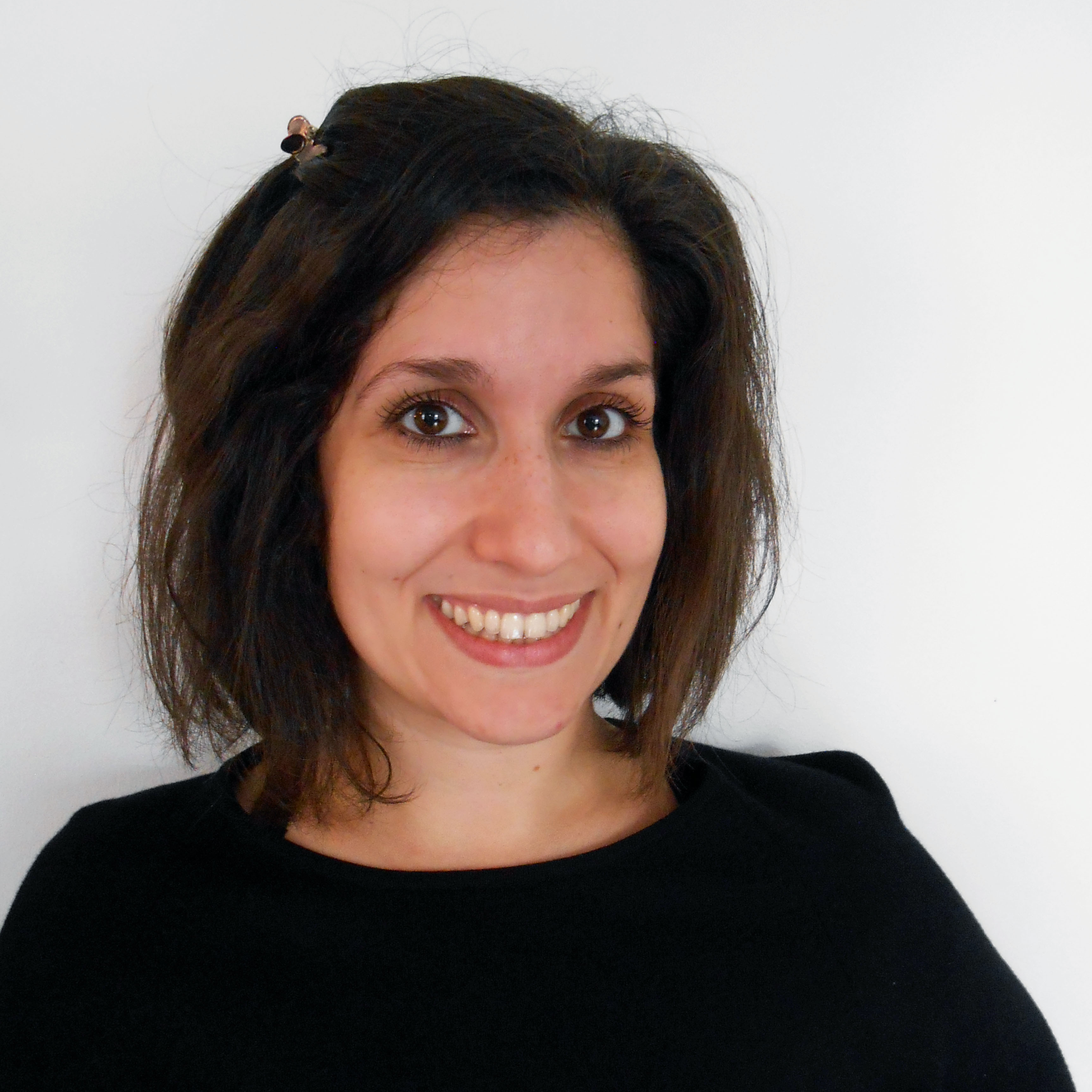 Headshot of a white female with chin length dark brown hair and dark brown eyes with a full smile. She is wearing a hair clip, plain long sleeved black shirt and is situated in front of a plain white background.