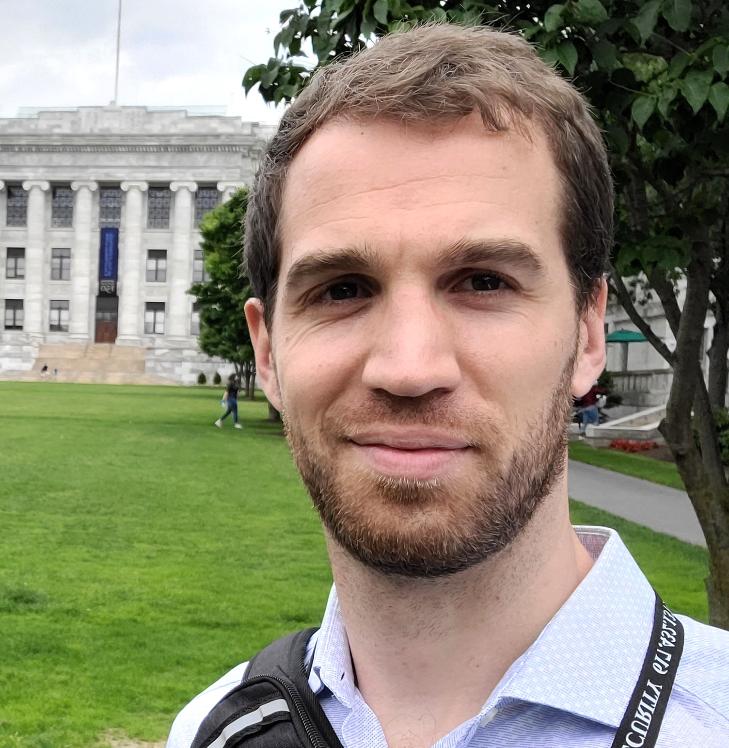 Headshot of a white male in his thirties on the lawn in front of the Harvard Medical School’s main building. He has brown eyes, short brown hair and a short beard and is slightly smiling. He’s wearing a blue button-up shirt with white specks.