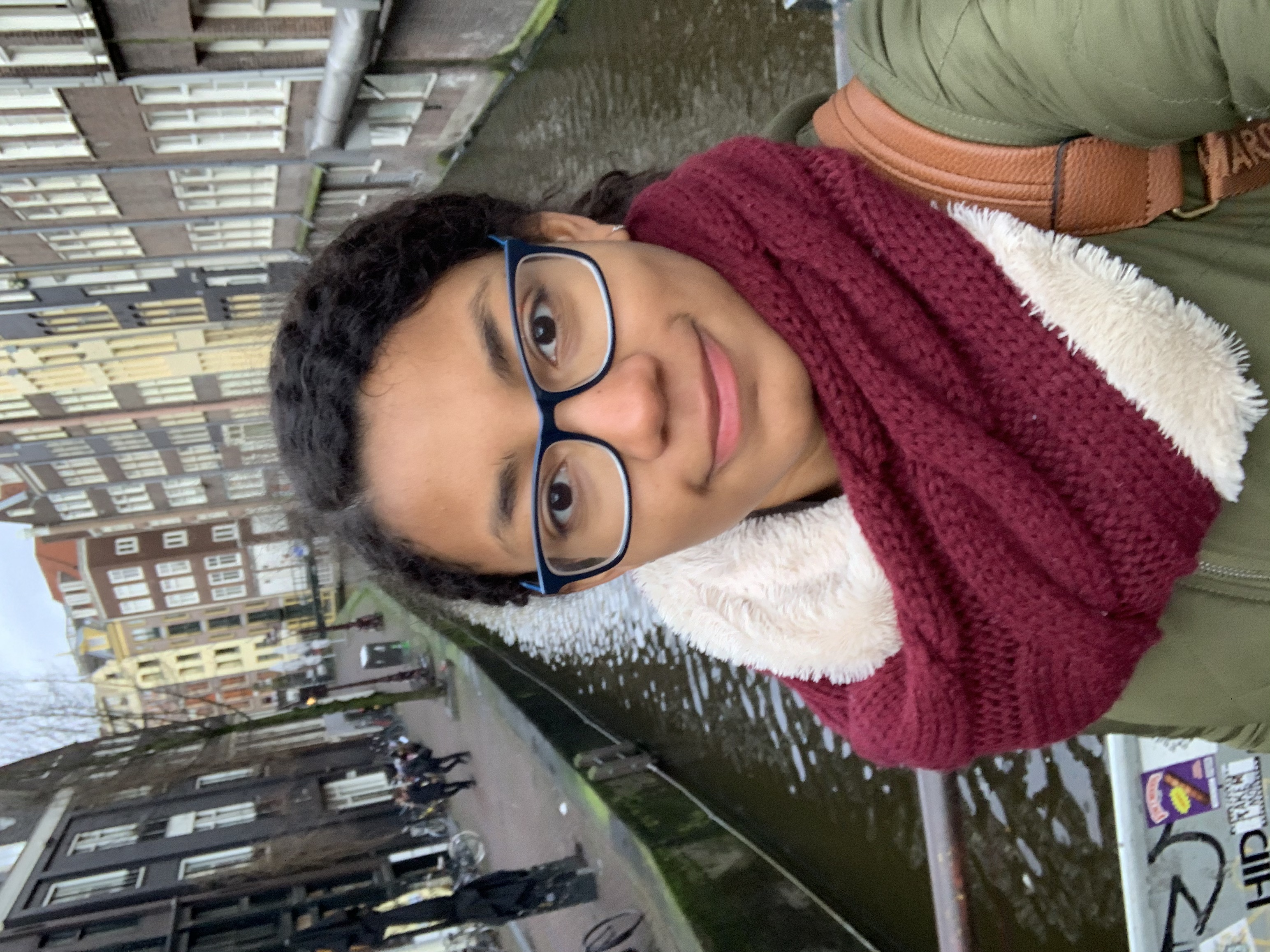 Selfie of a smiling young brown woman with dark hair and brown eyes. She is wearing rectangular glasses, a green jacket, and a maroon scarf. She is standing on a canal bridge in front of rows of brick buildings and bicycles.