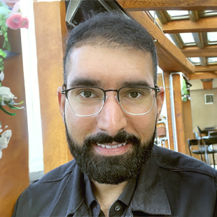 Headshot of an adult brown man with short black hair, a neatly trimmed full beard, and large, gold rimmed browline eyeglasses wearing a black button-up. He is situated in front of a blurred outdoor setting.