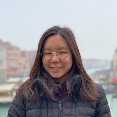 A headshot photo of a smiling young woman with long, straight, dark brown hair, wearing thin circular wire-rimmed glasses. The blurred background appears to be outdoors, featuring buildings and boats on both sides of a waterway.