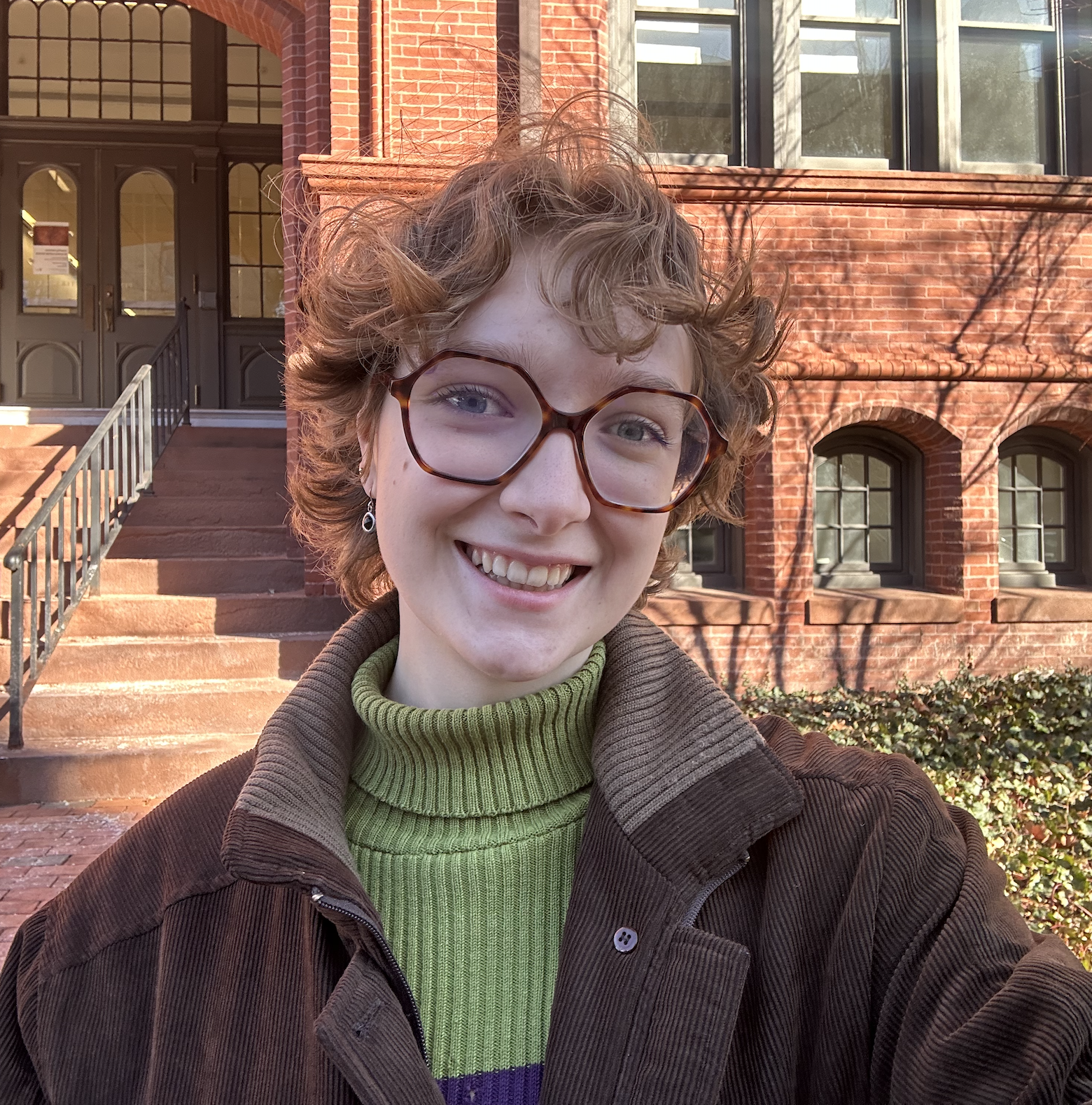 Blonde woman with hexagonal glasses smiles in front of a brick building.