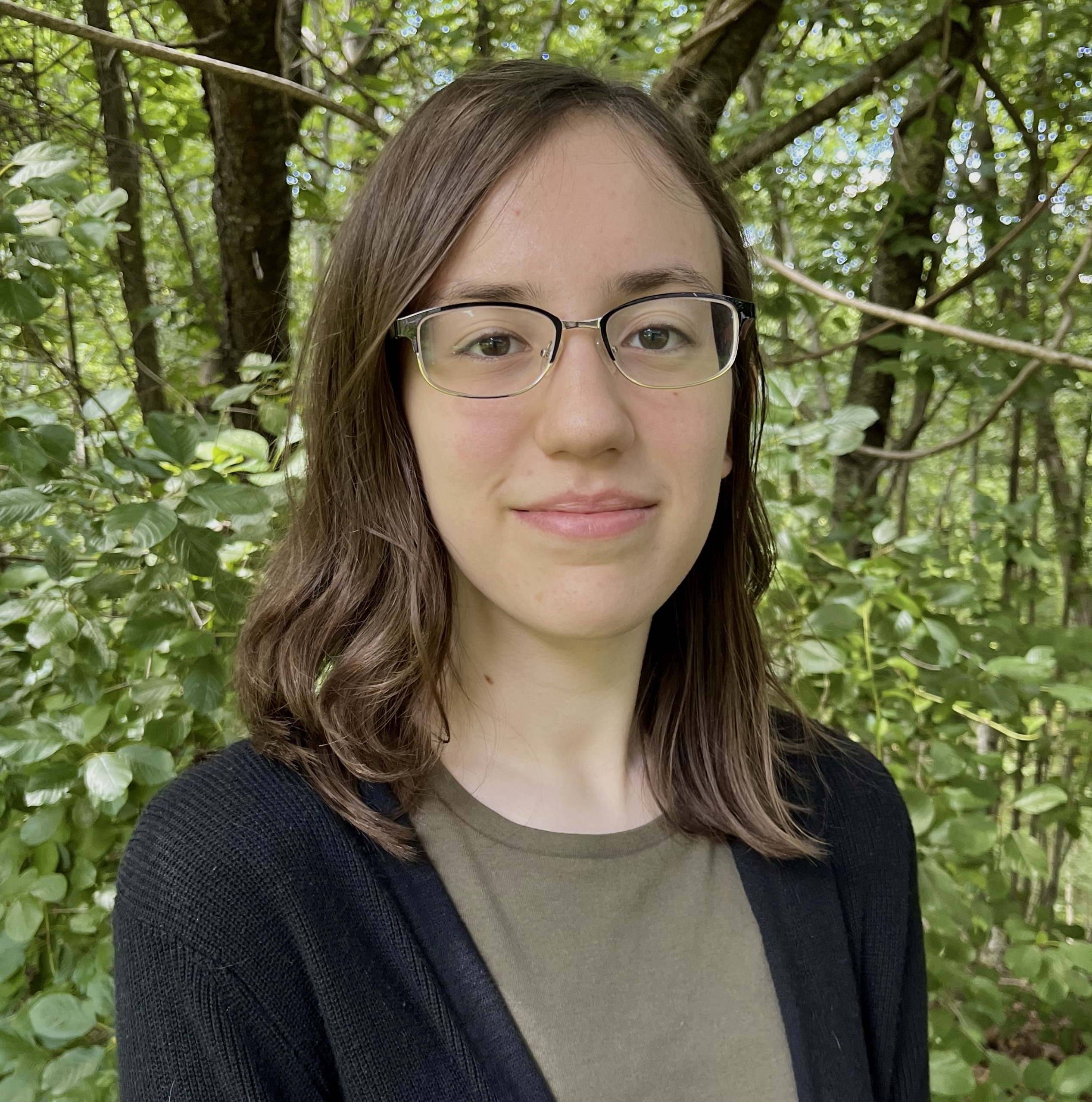 Photo of a smiling young woman with shoulder-length light brown hair and rectangular glasses. She is wearing an olive-colored shirt and a black cardigan, standing in the woods on a sunny day.