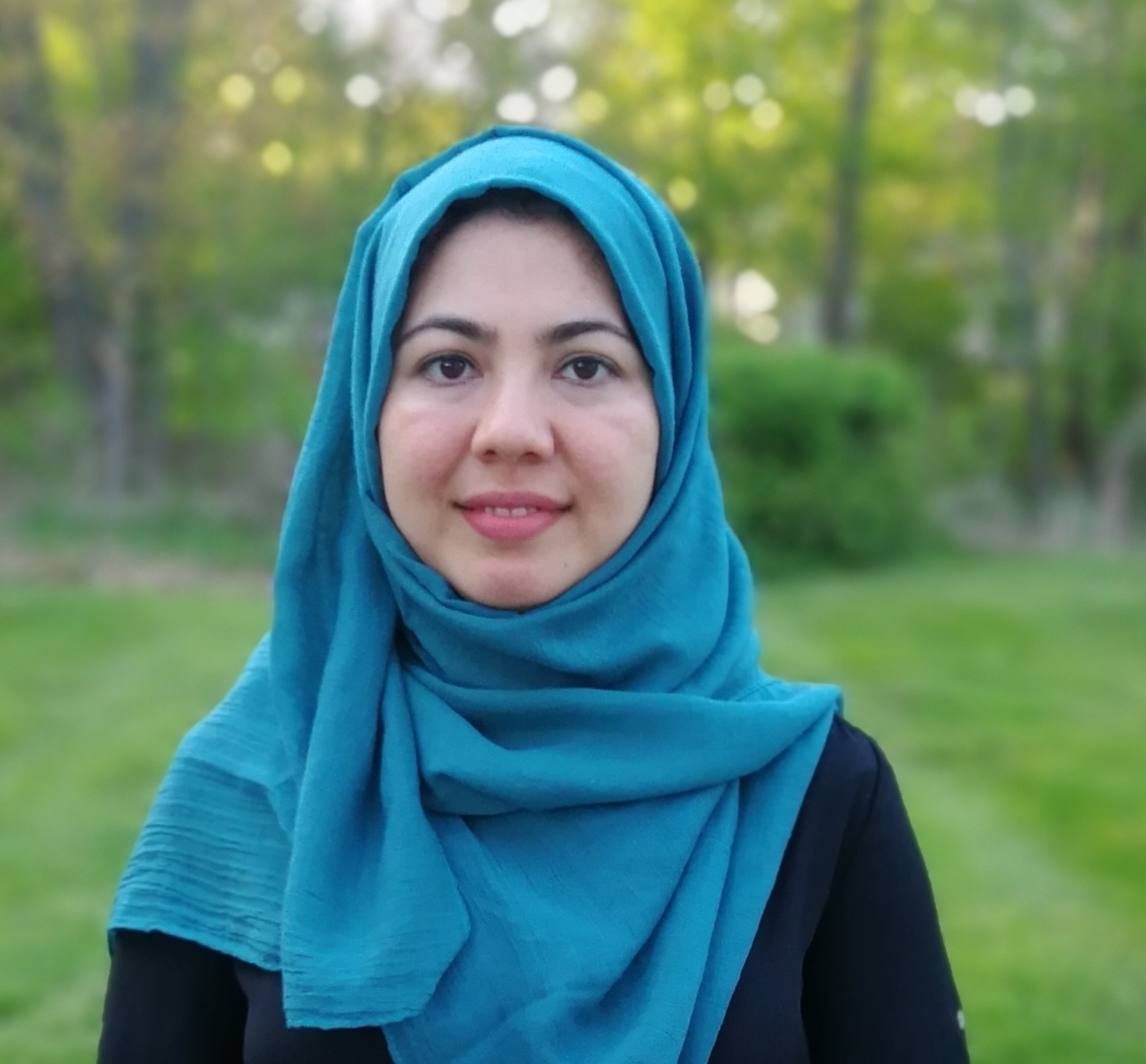 Photograph of a female wearing a blue scarf and a black shirt standing in what appears to be a park with trees and greenery.