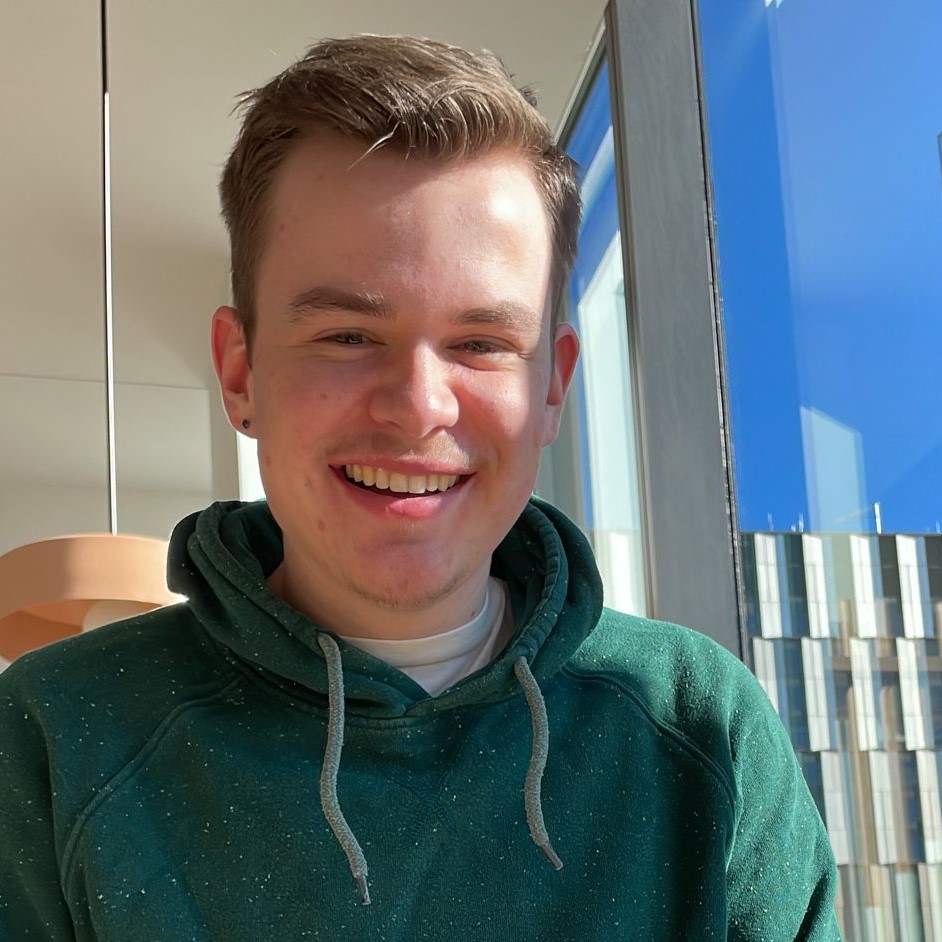 Photograph of a young white man with short blonde hair and a hint of stubble. He smiles wearing a green hoodie over a white tee shirt next to the windows inside of a brightly lit room.