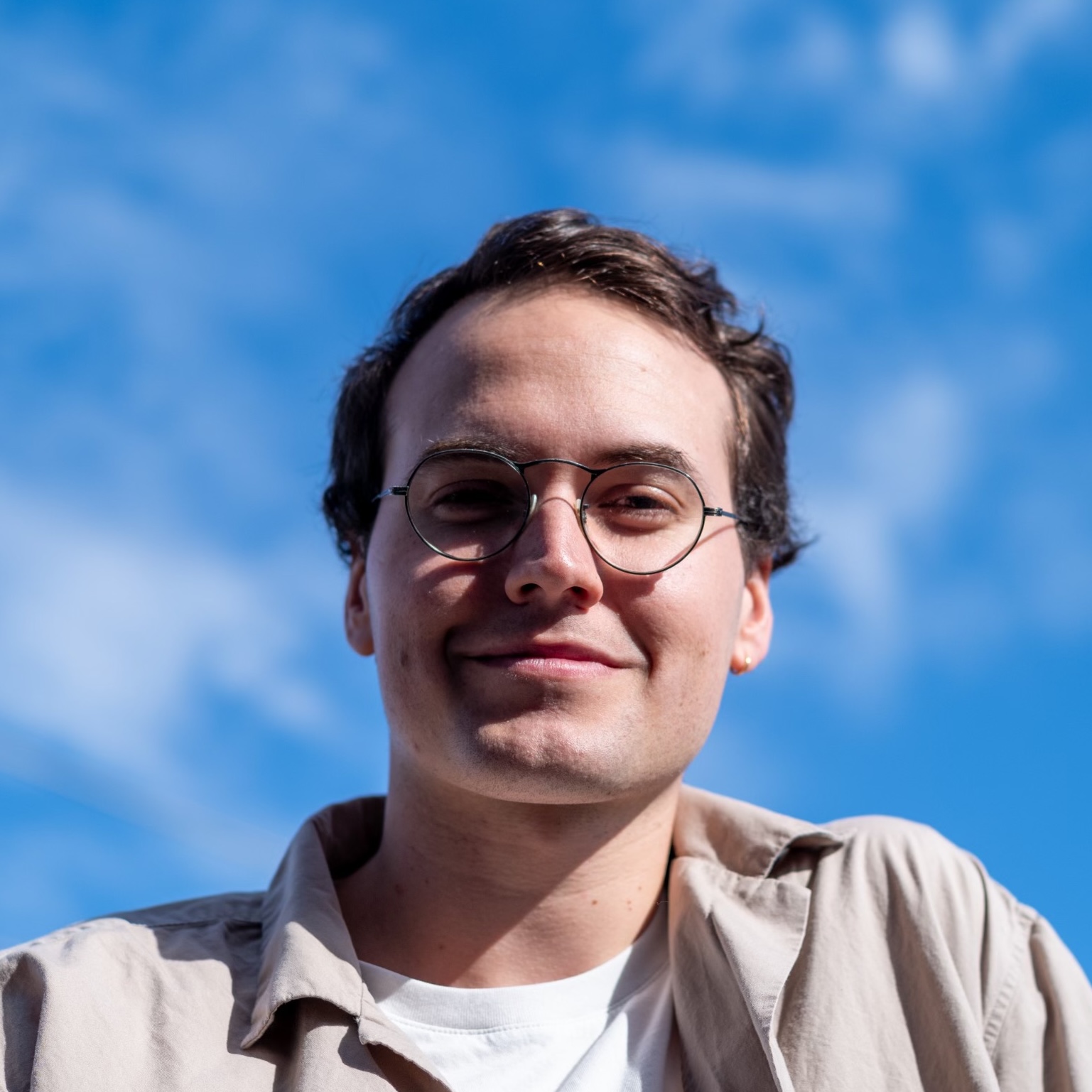 Headshot of a white male with a closed-mouth smile, wearing circular wire frame glasses. He has brown hair and the background is blurred blue sky.