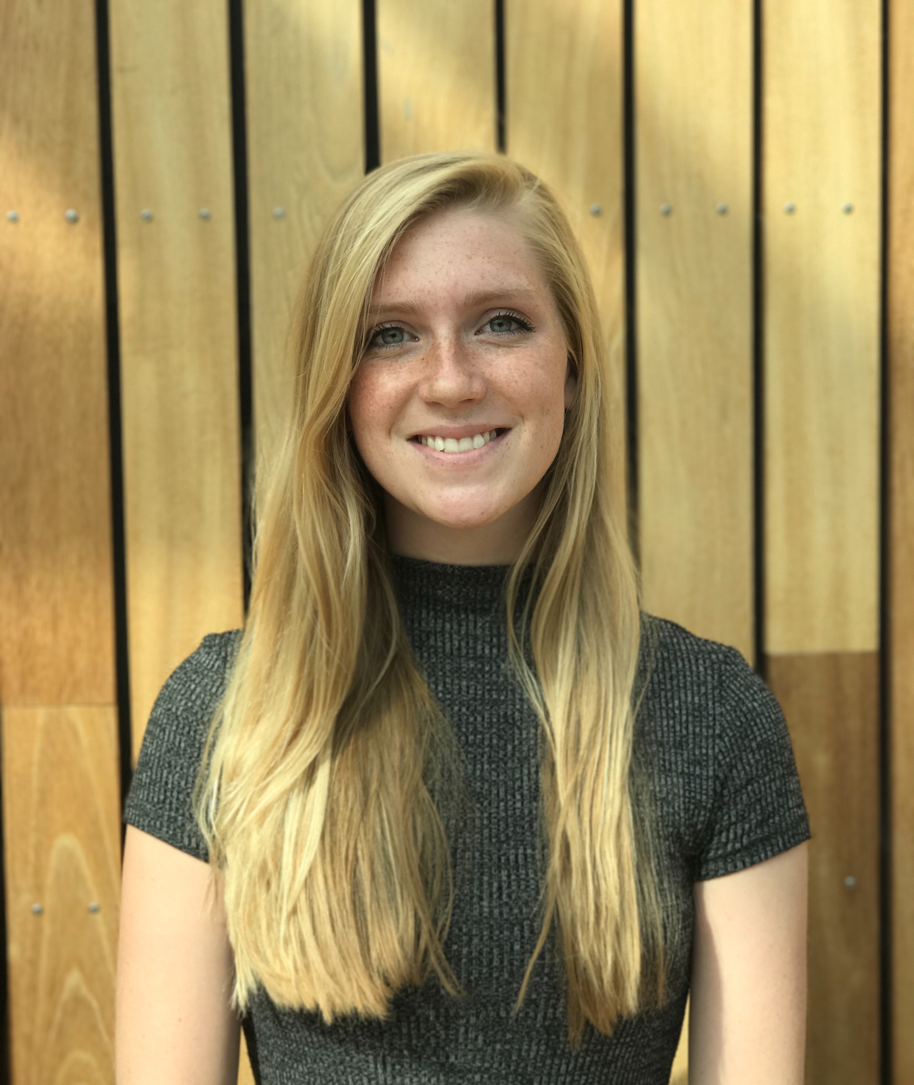 Photo of a young female who appears to be white, smiling, with long straight blonde hair. She is wearing a high-neck short sleeve shirt, in front of a light color wooden fence.