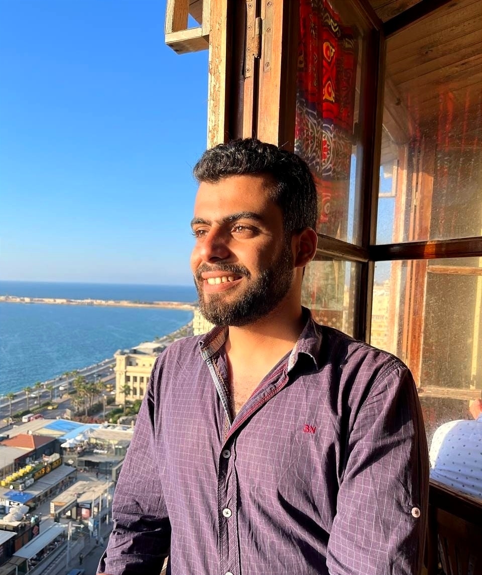 Photo of a young male who appears to be Asian with shortly cut dark curly hair and a beard. He’s wearing a purple button-up and smiling from a window into the distance. In the background, we can see the location is by a body of water.