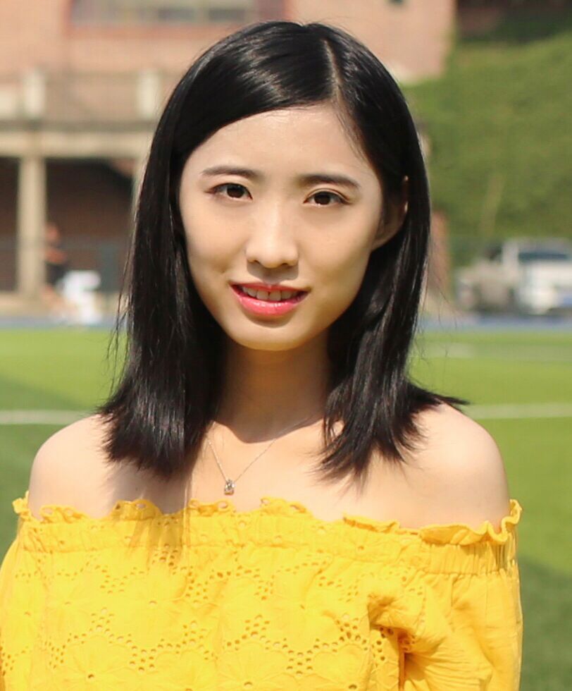 Headshot of a young woman who appears to be Asian, with medium-length black hair and an open smile, wearing a bright yellow top exposing her shoulders. The woman is outside in sunny weather, in front of a lawn with a blurry brick building in the background.