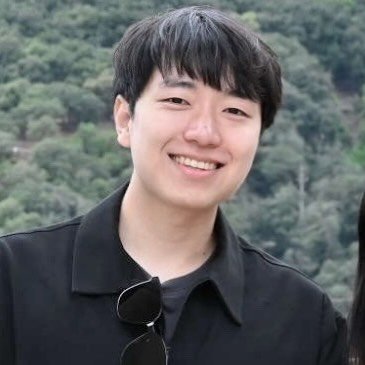 Outdoor portrait of a smiling young Asian man with short black hair, wearing a black jacket with sunglasses hanging from the collar, in front of a tree-covered hillside.