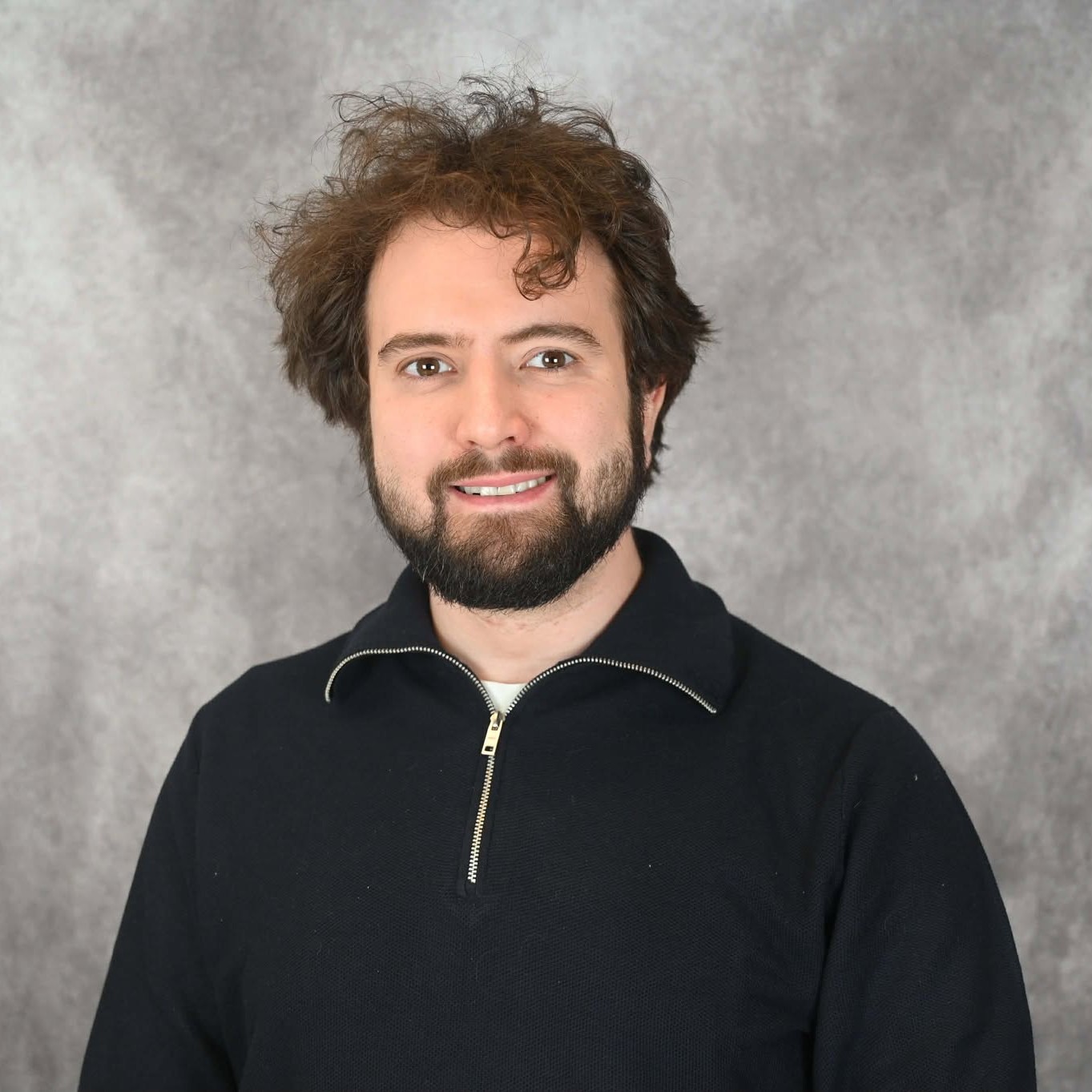 Smiling young white man with short curly brown hair and a beard, wearing a black shirt, in front of a blurred gray background.